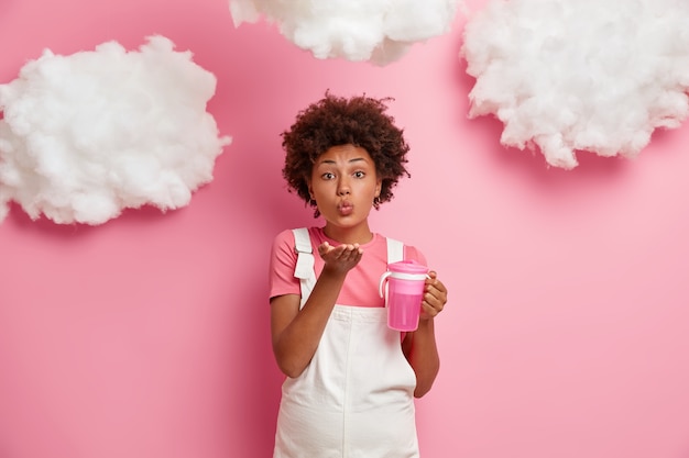 Hermosa mujer embarazada romántica espera bebé, sopla beso de aire al marido, viste ropa cómoda, sostiene la botella con agua fresca, aislada sobre una pared rosada con nubes esponjosas arriba. Maternidad