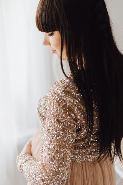 Hermosa mujer embarazada con vestido beige largo de pie en una habitación acogedora y luminosa