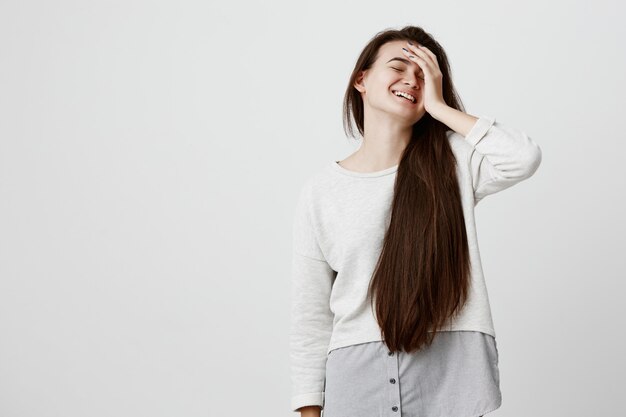 Hermosa mujer emocional con cabello largo y oscuro vestida casualmente, mantiene la mano sobre la cabeza, sonríe alegremente, ríe