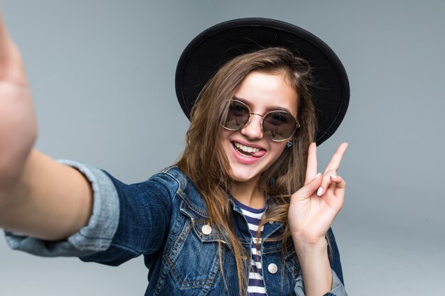 Hermosa mujer encantadora con gesto de paz con sombrero negro y gafas de sol tomar selfie de manos sobre fondo gris