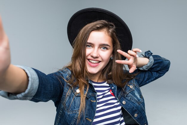 Hermosa mujer encantadora con gesto de paz con sombrero negro y gafas de sol tomar selfie de manos sobre fondo gris