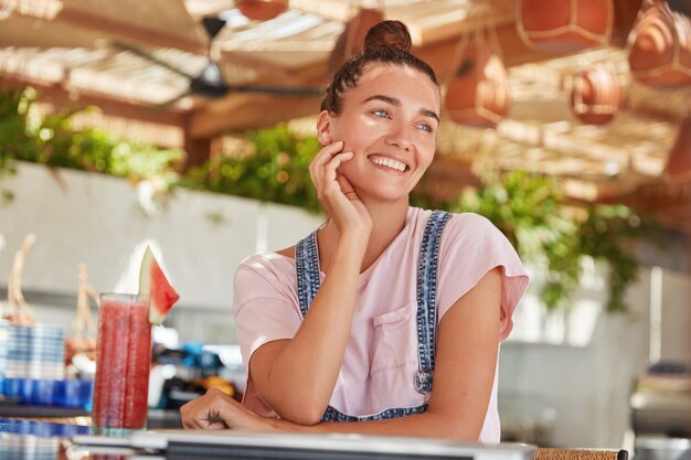 Hermosa mujer de ensueño con cálidos ojos azules, sonrisa positiva, vestida de manera informal, tiene el cabello oscuro atado en un nudo, bebe un cóctel fresco y le agrada pasar el tiempo libre en la cafetería. Descanso de verano, ocio