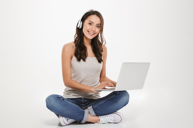 Hermosa mujer escuchando música o chateando con auriculares y una computadora portátil mientras está sentado con las piernas cruzadas en el piso, sobre la pared blanca