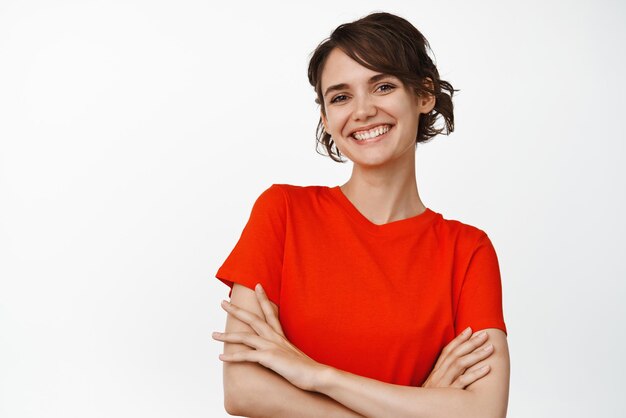 Hermosa mujer con estilo cruza los brazos en el pecho mirando confiada y sonriendo a la cámara de pie en camiseta roja contra el fondo blanco