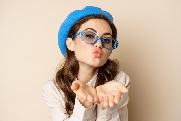 Hermosa mujer con estilo en gafas de sol soplando beso de aire en las manos cerca de los labios mirando suavemente y coqueta a...