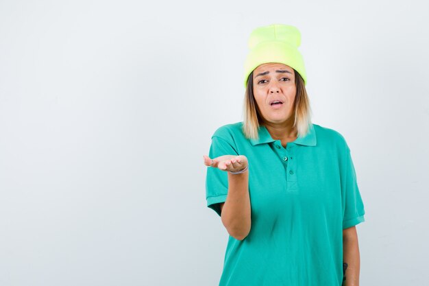 Hermosa mujer estirando la mano a la cámara en camiseta de polo, gorro y mirando desconcertado. vista frontal.