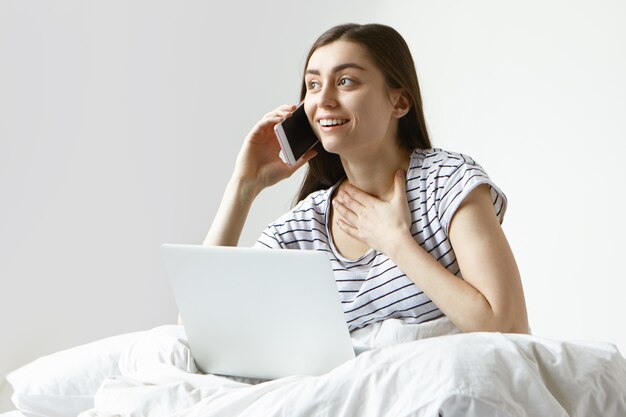 Hermosa mujer estudiante en pijama de rayas pasando la mañana en la cama, navegando por internet usando una computadora portátil y hablando por teléfono móvil