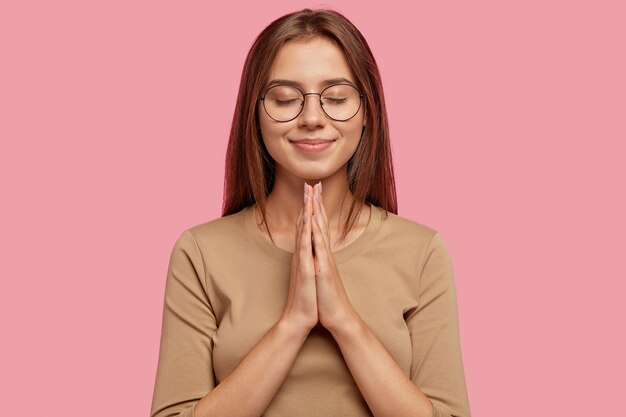 Hermosa mujer europea pide ayuda o pide disculpas mientras mantiene las manos en gesto de oración, usa anteojos redondos, tiene cabello oscuro, vestida con ropa informal, aislada sobre una pared rosa