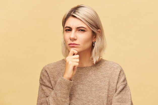 Hermosa mujer europea sospechosa joven de moda en jersey de cachemira sosteniendo la mano en la barbilla y mirando con sospecha y desconfianza, arrugando los ojos. Expresiones faciales humanas