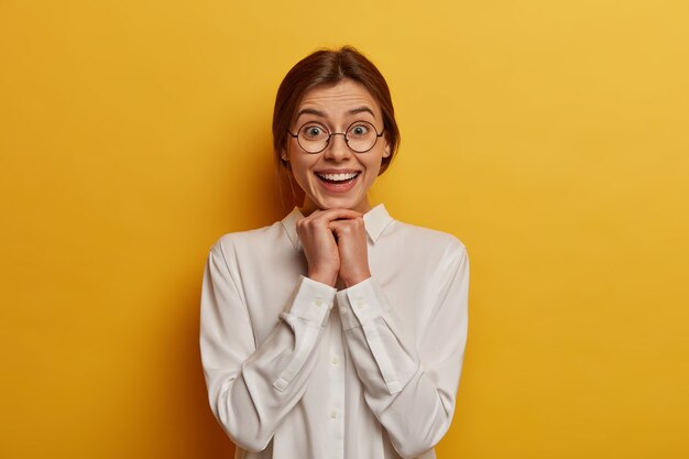 Hermosa mujer con expresión alegre, mantiene las manos debajo de la barbilla, viste una pulcra camisa blanca y grandes anteojos redondos, se ve alegre, de buen humor, aislada sobre una pared amarilla.