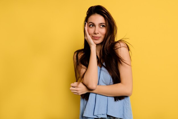 Hermosa mujer feliz en camisa azul que cubre la cara con la mano y mira soñadora en el marco de la pared amarilla.