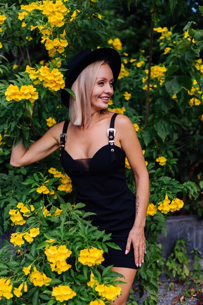 Hermosa mujer feliz caucásica con estilo en vestido negro y sombrero clásico en el parque rodeado de flores amarillas tailandesas