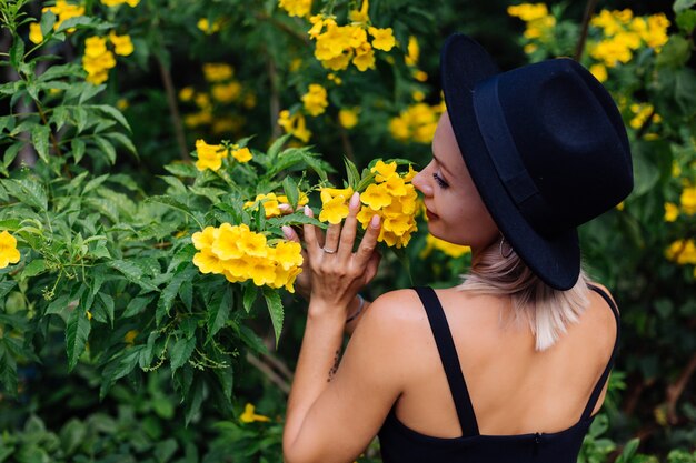 Hermosa mujer feliz caucásica con estilo en vestido negro y sombrero clásico en el parque rodeado de flores amarillas tailandesas