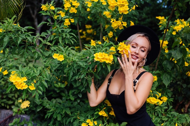 Hermosa mujer feliz caucásica con estilo en vestido negro y sombrero clásico en el parque rodeado de flores amarillas tailandesas