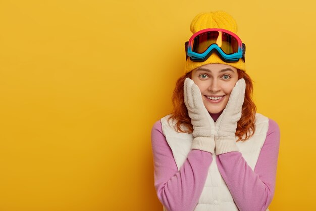 Hermosa mujer feliz mantiene ambas manos en las mejillas, tiene una sonrisa tierna, mira con una sonrisa a la cámara, disfruta del descanso activo y del esquí de travesía, vestida con ropa deportiva, aislada en la pared amarilla. Horario de invierno.