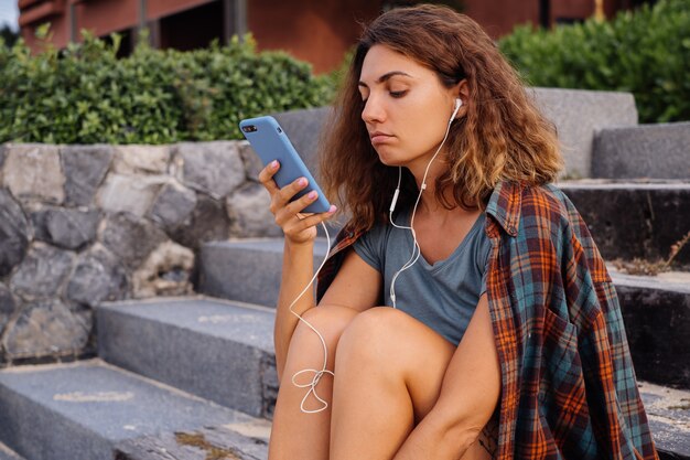 Hermosa mujer en forma en pantalones cortos de jean, camisa a cuadros sentarse en las escaleras a la luz del atardecer con smartphone