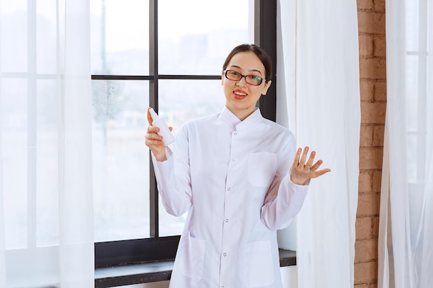 Hermosa mujer con gafas en bata de laboratorio con desinfectante en aerosol cerca de la ventana.