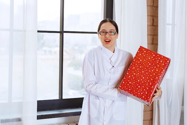 Hermosa mujer con gafas en bata de laboratorio con gran caja de regalo junto a la ventana.