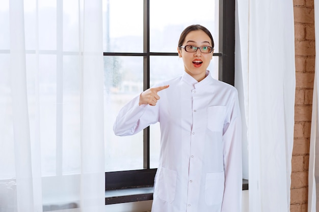 Hermosa mujer con gafas y bata de laboratorio de pie junto a la ventana y apuntando a un lado.