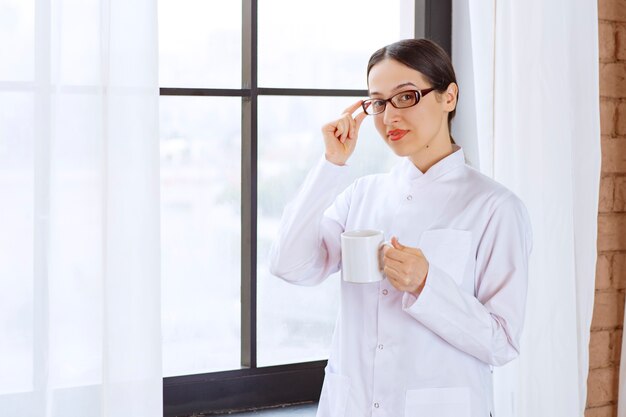 Hermosa mujer con gafas en bata de laboratorio de pie mientras sostiene una taza de café junto a la ventana.