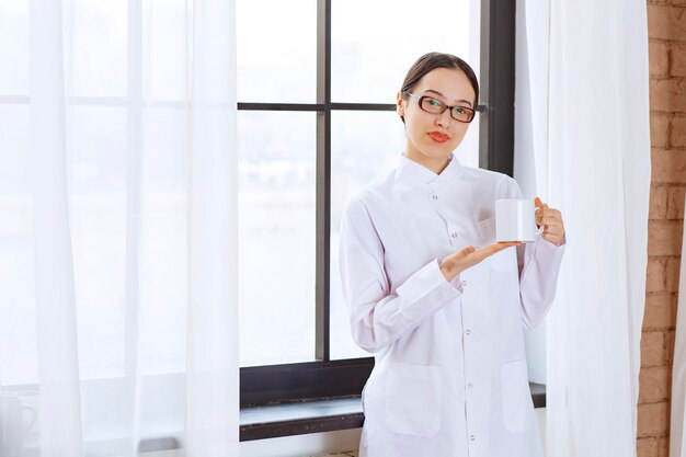 Hermosa mujer con gafas en bata de laboratorio posando con una taza de café junto a la ventana.