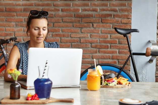 Hermosa mujer con gafas de sol en la cabeza, navegando por Internet, revisando su suministro de noticias a través de las redes sociales y enviando mensajes en línea, usando wifi gratis en la cafetería moderna