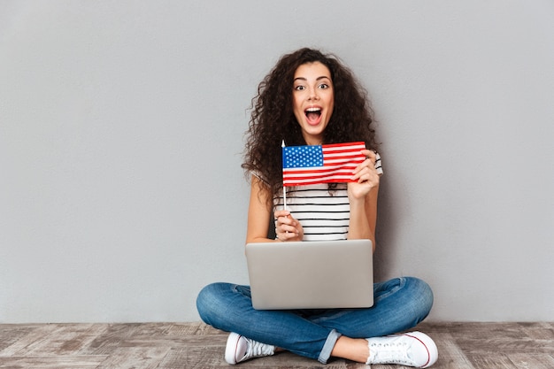 Hermosa mujer con hermosa sonrisa sentada en postura de loto con computadora plateada en las piernas mostrando la bandera americana sobre la pared gris