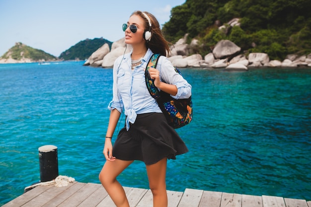 Hermosa mujer hipster viajando alrededor del mundo con mochila, sonriendo, feliz, positivo, escuchando música en auriculares, fondo azul océano tropical, gafas de sol, sexy, vacaciones de verano,