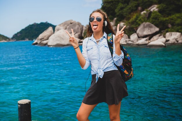 Foto gratuita hermosa mujer hipster viajando alrededor del mundo con mochila, sonriendo, feliz, positivo, escuchando música en auriculares, fondo azul océano tropical, gafas de sol, sexy, vacaciones de verano,