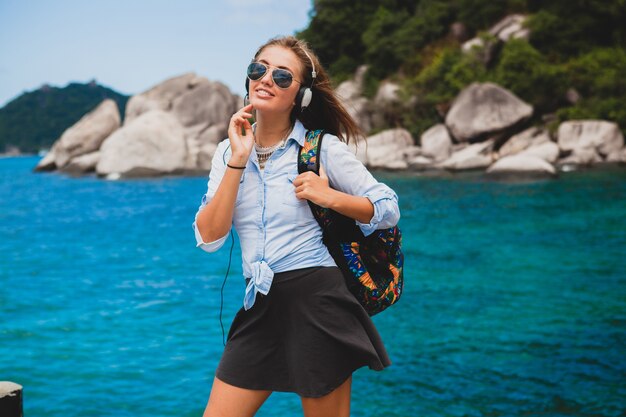 Hermosa mujer hipster viajando alrededor del mundo con mochila, sonriendo, feliz, positivo, escuchando música en auriculares, fondo azul océano tropical, gafas de sol, sexy, vacaciones de verano,