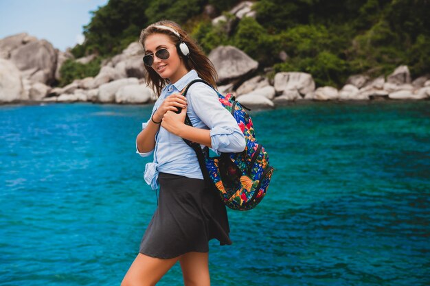 Hermosa mujer hipster viajando alrededor del mundo con mochila, sonriendo, feliz, positivo, escuchando música en auriculares, fondo azul océano tropical, gafas de sol, sexy, vacaciones de verano,