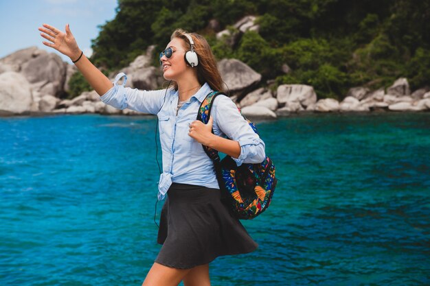 Hermosa mujer hipster viajando alrededor del mundo con mochila, sonriendo, feliz, positivo, escuchando música en auriculares, fondo azul océano tropical, gafas de sol, sexy, vacaciones de verano,