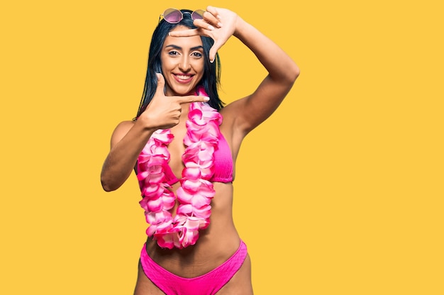 Hermosa mujer hispana con bikini y lei hawaiano sonriendo haciendo marco con manos y dedos con cara feliz creatividad y concepto de fotografía