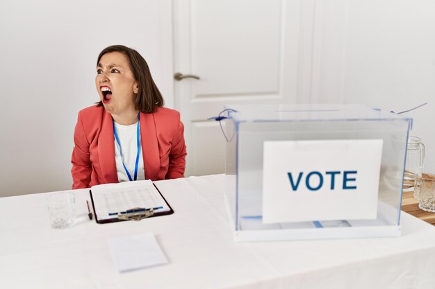 Hermosa mujer hispana de mediana edad en las elecciones políticas sentada junto a la boleta enojada y enojada gritando frustrada y furiosa, gritando con ira. rabia y concepto agresivo.