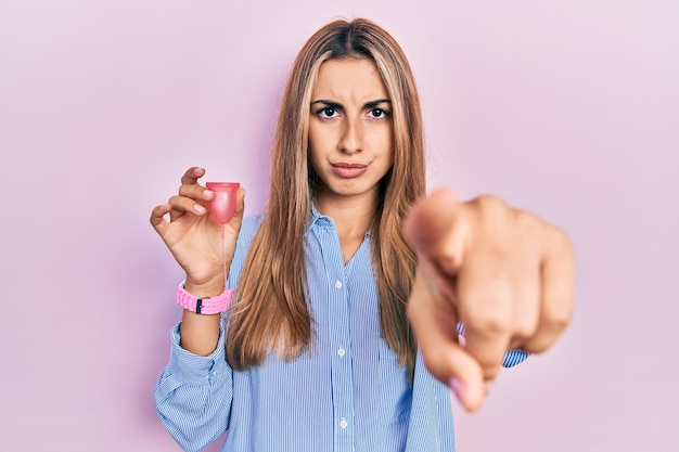 Hermosa mujer hispana sosteniendo copa menstrual apuntando con el dedo a la cámara y a ti, gesto de confianza que parece serio