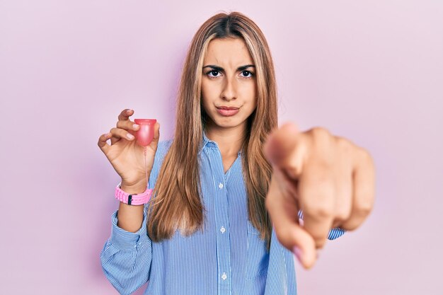 Hermosa mujer hispana sosteniendo copa menstrual apuntando con el dedo a la cámara y a ti, gesto de confianza que parece serio