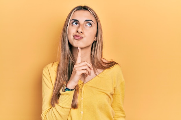 Hermosa mujer hispana con suéter amarillo casual pensando concentrada en la duda con el dedo en la barbilla y mirando hacia arriba preguntándose