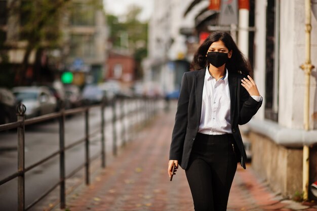 Hermosa mujer india usa una máscara formal y negra posando en la calle durante la pandemia de covid con el teléfono celular a mano