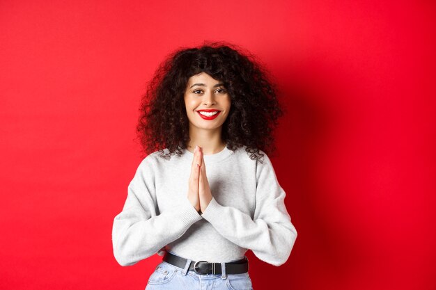 Hermosa mujer italiana sonriente diciendo gracias, tomados de la mano en gesto namaste y mirando a la cámara agradecida, de pie sobre fondo rojo.