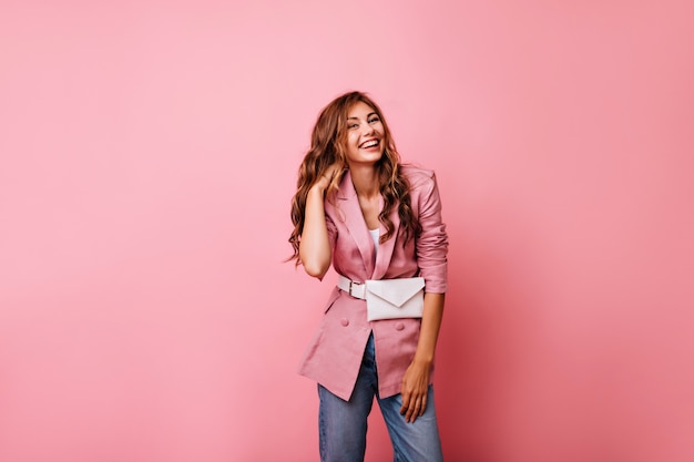 Hermosa mujer de jengibre en chaqueta casual riendo en rosa. Señora joven soñadora en jeans jugando con su pelo rojo rizado.