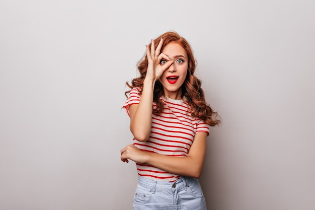 Hermosa mujer de jengibre rizado disfrutando de la sesión de fotos en interiores. Chica caucásica extática de moda peinado casual de pie en la pared blanca.