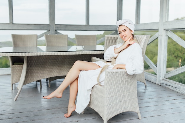 Hermosa mujer joven en bata blanca, sentada en la terraza de verano en su casa. Concepto de mañana.