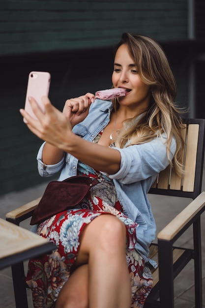 Hermosa mujer joven con cabello largo come helado y hace selfie en smartphone. retrato de primer plano afuera