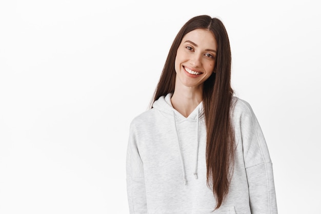 Hermosa mujer joven con cabello largo y oscuro, inclina la cabeza y sonríe al frente con ternura, de pie franca y positiva contra la pared blanca, vistiendo una sudadera con capucha