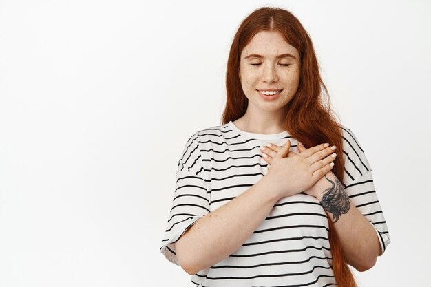 Hermosa mujer joven con cabello largo rojo y piel pálida, tiene recuerdos, soñando despierto, tomados de la mano en el corazón y sonriendo con los ojos cerrados, recuerda algo en blanco