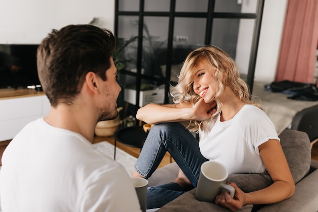 Hermosa mujer joven con cabello rubio y taza mirando a su novio y sonriendo. Feliz pareja sentada en casa en el sofá.