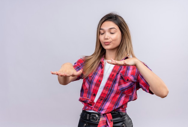 Foto gratuita una hermosa mujer joven en camisa a cuadros de pie con los ojos cerrados y tratar de tocar algo o encontrar en una pared blanca