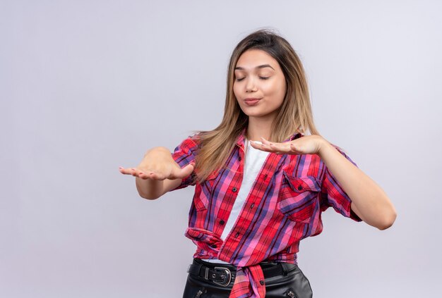 Una hermosa mujer joven en camisa a cuadros de pie con los ojos cerrados y tratar de tocar algo o encontrar en una pared blanca