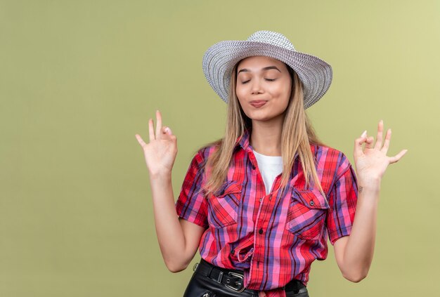 Una hermosa mujer joven con una camisa a cuadros con sombrero cerrando los ojos mientras muestra un gesto de ok en una pared verde