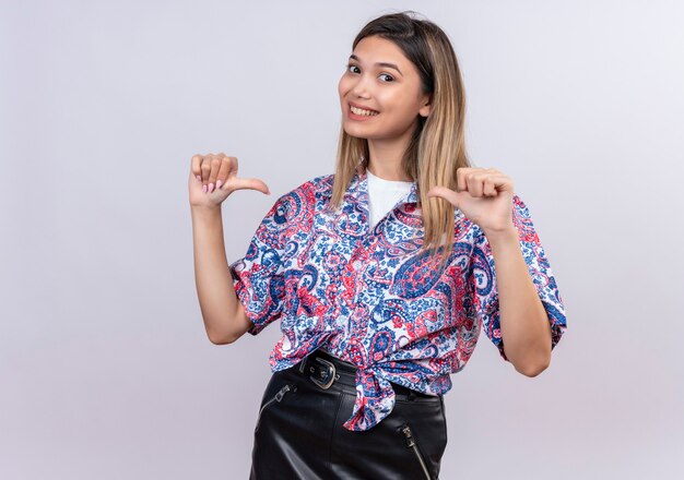 Una hermosa mujer joven con camisa estampada de paisley apuntando con el pulgar a sí misma mientras mira en una pared blanca